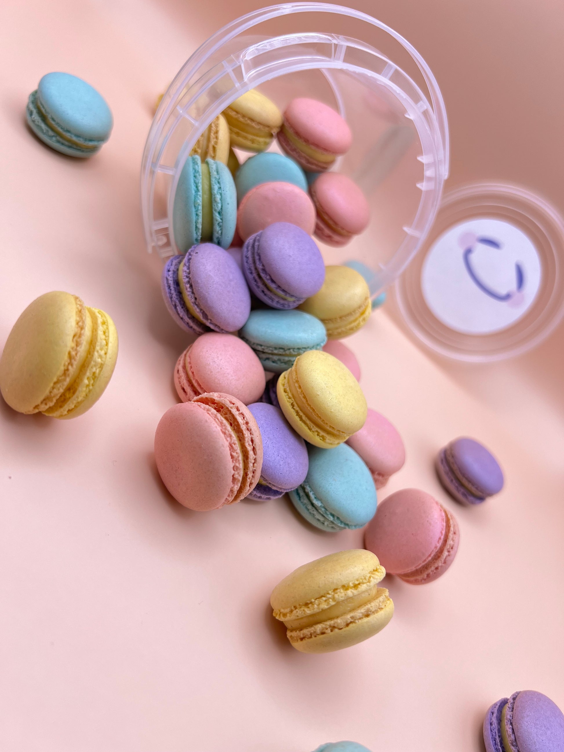 Pastel-colored mini macarons spilling out of a clear container on a pink background